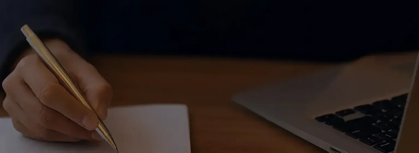hero picture home page A hand holding a pen above a notebook, with a laptop partially visible.